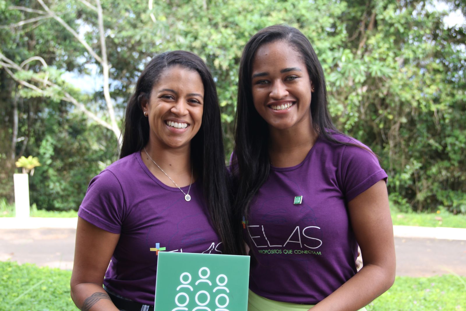 Duas mulheres negras sorriem com uma placa representando ações sociais